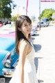 A woman in a white dress standing in front of a blue car.